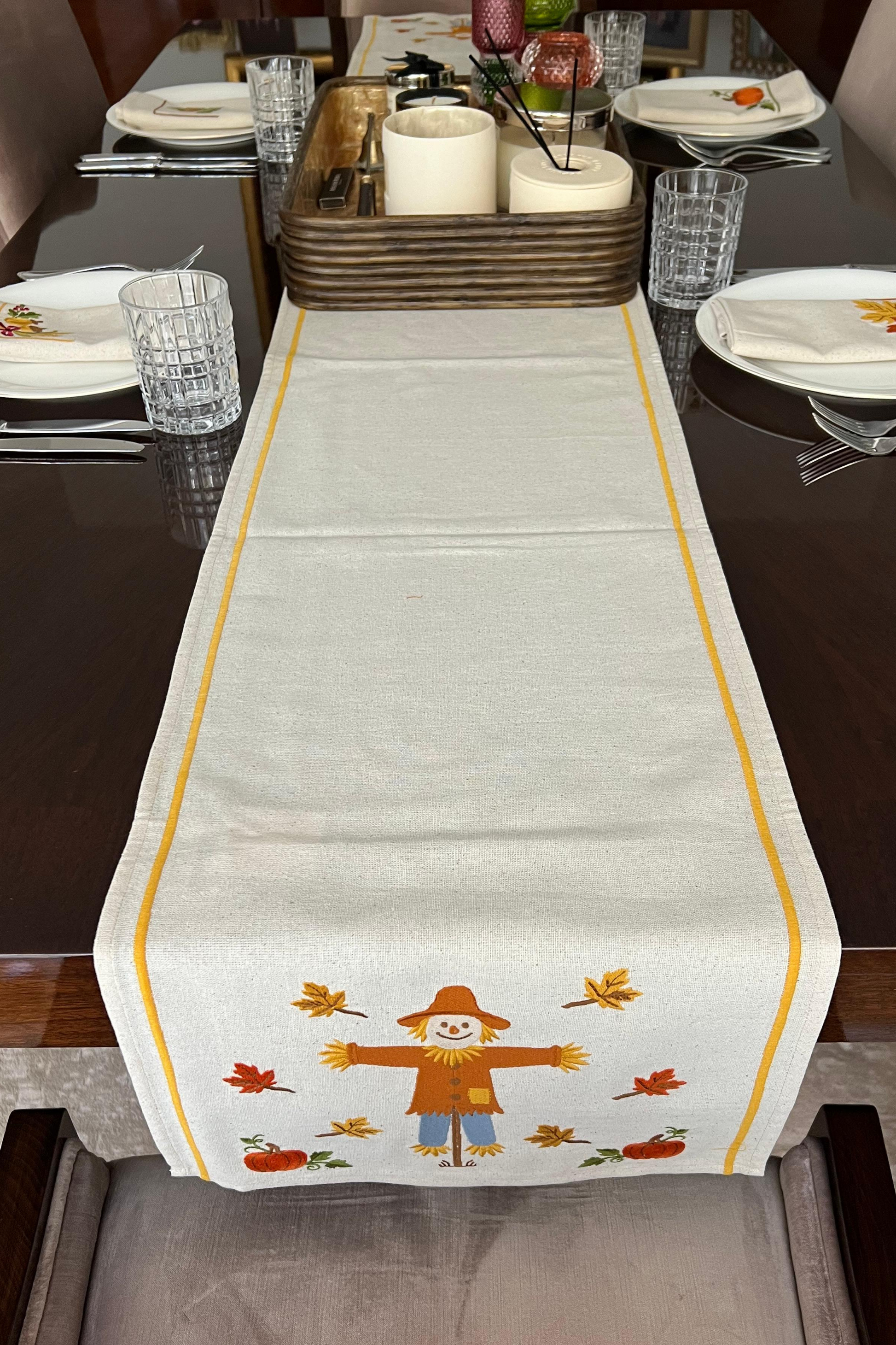 Table setting with a decorative table runner and napkins featuring a scarecrow, pumpkins, and autumn leaves.