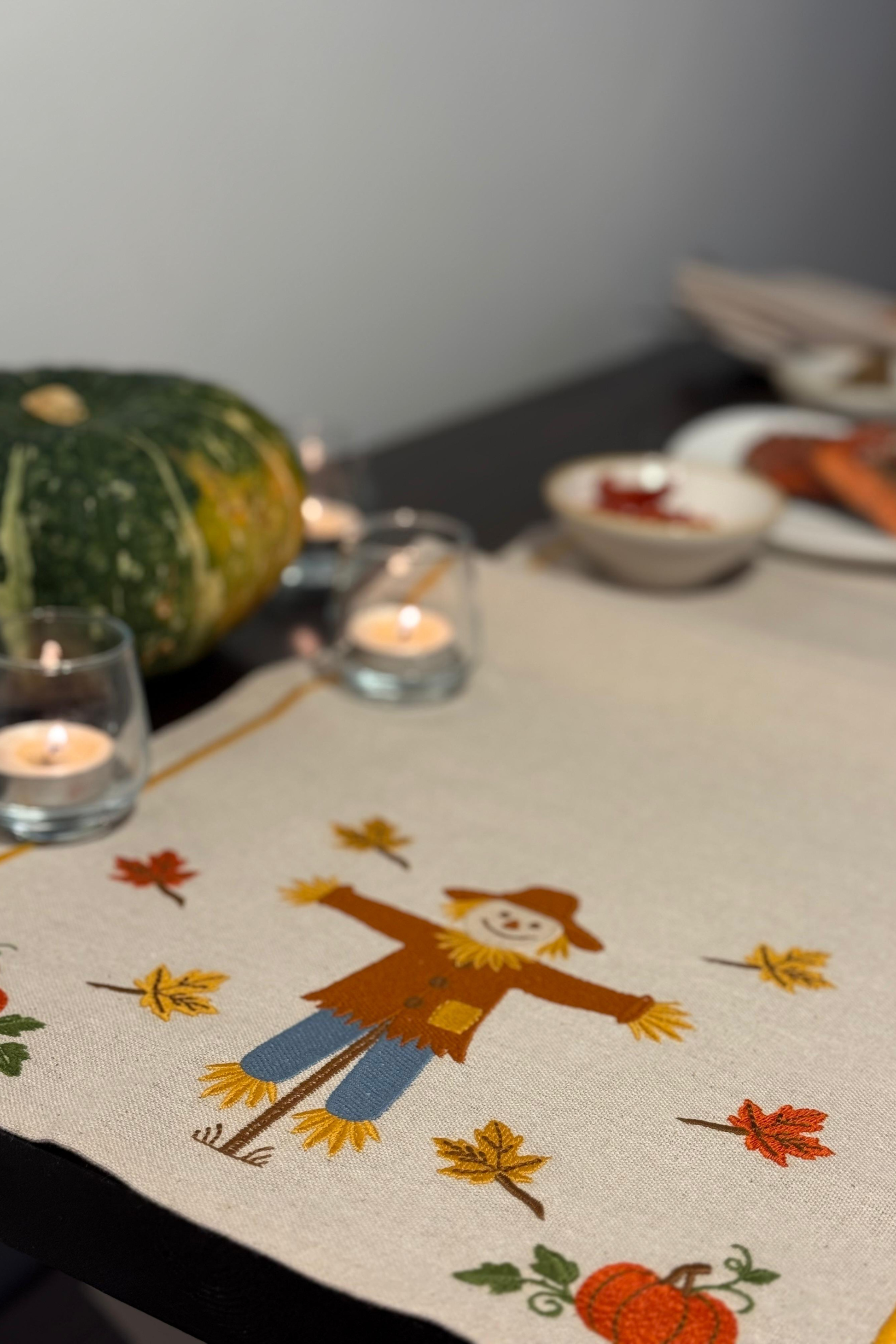 Table setting with a table runner featuring a scarecrow and autumn elements, candles, and a pumpkin.